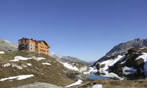 Das Pfitscher Joch Haus thront auf einem Hügel neben einem Bergsee. Die Alpenlandschaft ist von Schnee und braunem Gras bedeckt.