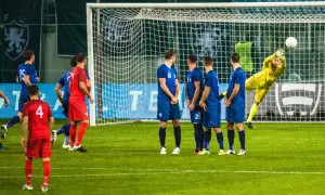 Piłkarze w niebieskich strojach czekają na wznowienie gry. Bramkarz w żółtym stroju broni strzał. Czas na przerwę w meczu?