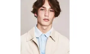 Ragazzo con capelli castani mossi e acconciature alla moda, indossa una camicia azzurra e una giacca beige.