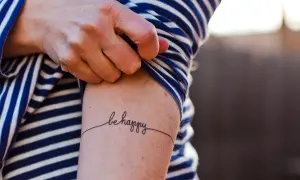 Un brazo con una camiseta a rayas azules y blancas muestra un delicado tatuaje de caligrafía que dice "be happy", una forma de arte para transformar cicatrices.
