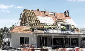 Ekipa budowlana pracuje nad dachem domu. Widać rusztowania, drabiny i materiały budowlane. To pokazuje, jaki jest koszt dachu.