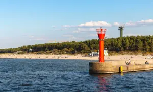 Mrzeżyno atrakcje: plaża, morze i charakterystyczna czerwona wieża nawigacyjna na falochronie. W tle las i zielona wieża.