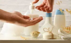 Hands dispensing lotion from a pump bottle into a palm, with oat flakes, soap, and bottles in the background.