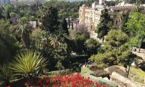 Vista panorámica de un jardín exuberante con flores rojas y palmeras, un edificio histórico y el mar al fondo en Málaga.