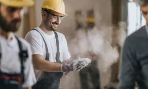 Budowlaniec w żółtym kasku i goglach uśmiecha się, otrzepując rękawice z pyłu. W tle widać innych pracowników, a w powietrzu unosi się pył, być może pochodzący z materiałów zawierających azbest.