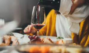 Rödvinet hälls upp i ett glas vid en middagsbjudning. En person i gul tröja sitter bakom. Frågan om hur mycket vin per person är relevant vid sådana tillfällen.
