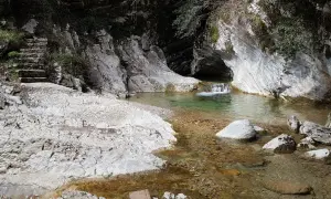 Sentiero di pietra conduce a un ruscello cristallino con piccole cascate di piovere, incastonato tra rocce bianche e vegetazione.