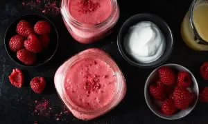 Två glas med en rosa, nyttig smoothie toppad med frystorkade hallon. Bredvid finns skålar med färska hallon och yoghurt, samt ett glas juice.