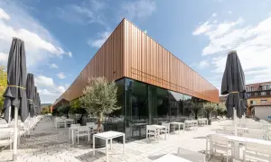 Moderne Architektur der Inselhalle Lindau mit kupferfarbener Fassade, Glasfront und vielen leeren Tischen und Stühlen auf einer gepflasterten Terrasse.