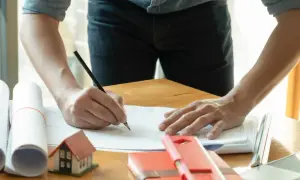 Architekt pisze ołówkiem na planach, obok makieta domu i zwój dokumentów.