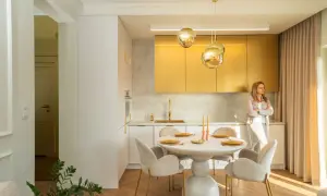Elegancka kuchnia z błyszczącymi, złotymi frontami szafek. Okrągły stół z krzesłami w pastelowych kolorach.