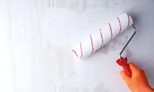 Ręka w pomarańczowej rękawicy wykonuje gruntowanie przed malowaniem, używając wałka z czerwonymi paskami.