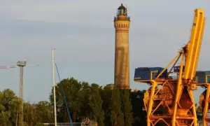 Wysoka, żółta latarnia morska w Gąskach góruje nad portem, obok potężnych, żółtych dźwigów portowych i zielonych drzew.