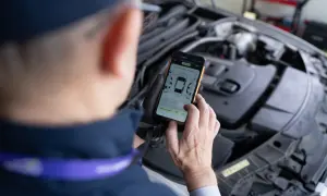 Mechanik w rękawiczkach trzyma smartfon z aplikacją do oceny stanu samochodu, na tle otwartej maski silnika.