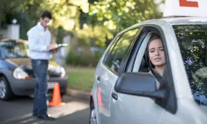 Joven nerviosa al volante, con un instructor anotando. ¿Se puede sacar el carnet de conducir sin autoescuela?