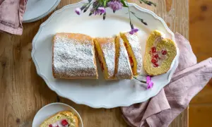 Eine köstliche Erdbeer Biskuitrolle, mit Puderzucker bestäubt und mit frischen Erdbeeren gefüllt, liegt auf einer weißen Platte.