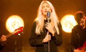 Wokalistka z blond włosami śpiewa przy mikrofonie, w tle zespół z gitarami i światła.