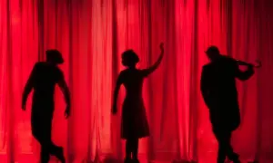 Sylwetki aktorów na tle czerwonej kurtyny. W centrum kobieta z uniesioną ręką, po bokach mężczyźni. Może to scena z przedstawienia założyciela Teatru Kamienica.
