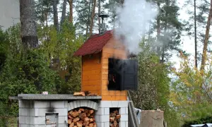 Gotowa budowa wędzarni ogrodowej. Dym unosi się z komina, a w środku wiszą kiełbaski.