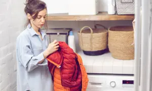 Frau prüft einen Daunenmantel, bereit zum wollmantel waschen. Daneben stehen Wäschekörbe und eine Waschmaschine.