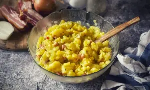 Ein dampfender, warmer Kartoffelsalat mit Speck und Zwiebeln, serviert in einer Glasschüssel mit Holzlöffel.