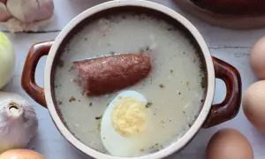 Gotowy zakwas na żurek śląski rączka gotuje. W garnuszku pływa kiełbasa i jajko. Obok cebula, czosnek i jajka.