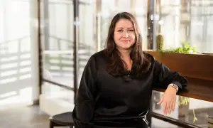 Aida Alvinius, en kvinna med långt brunt hår, ler mot kameran. Hon bär en svart klänning och sitter vid ett träbord med växter i bakgrunden.