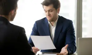 Mężczyzna w garniturze z niezadowoloną miną trzyma dokument, gestykulując ręką.