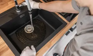 Wasser fließt in die Spüle, der Abfluss gluckert aber nicht verstopft. Ein Mann mit Handschuhen prüft den Wasserfluss.