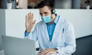 Lekarz w maseczce i kitlu wita pacjenta podczas konsultacji telemedycznej.