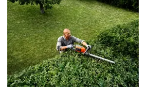 Hombre con gafas y guantes realiza la poda de setos con una cortasetos eléctrica. El césped verde y un árbol completan la escena.
