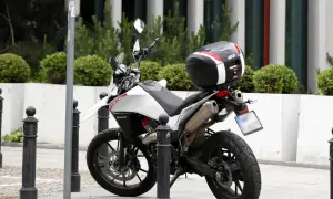 Biały motocykl z czarnym kufrem Givi zaparkowany na chodniku, obok słupków.