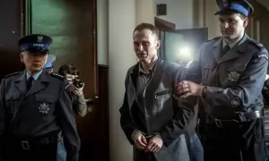 Scena z filmu o Tomaszu Komendzie: policjanci eskortują go w kajdankach, a za nimi stoi fotograf z aparatem.