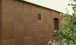 Brązowy cokół budynku, wyłożony płytkami o nieregularnym układzie, z małym okienkiem i kratką wentylacyjną.