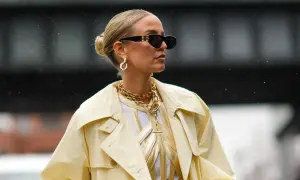Kobieta w okularach przeciwsłonecznych i żółtym płaszczu, z eleganckim upięciem włosów w kok. Złota biżuteria dodaje szyku.