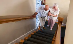 Coppia anziana scende le scale con fatica. Lui si appoggia al corrimano, lei gli tiene la mano.