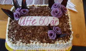 En tårtbotten i långpanna dekorerad med chokladspån, lila rosor och texten "Uffe 60 år".