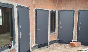 Nowe, szare drzwi zewnętrzne i okno w budowie. Widać ceglane ściany i materiały budowlane.