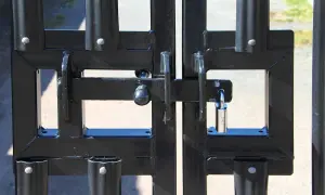 Zamykanie bramy przesuwnej. Solidna konstrukcja z czarnego metalu, zabezpieczona kłódką.