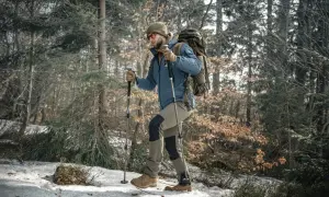 Mężczyzna w czapce i okularach przeciwsłonecznych idzie przez zaśnieżony las, wspierając się na kijach trekkingowych.