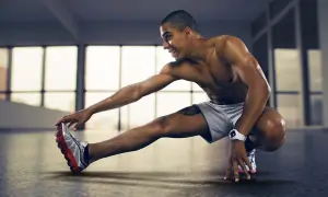 Ein Mann dehnt seine Beinbeuger in einem hellen Raum. Er ist nackt bis auf eine graue Shorts und Turnschuhe.