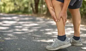 Uomo con dolore alle gambe, difficoltà a camminare su un sentiero ombreggiato.