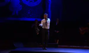Un bailaor flamenco español actual, con camisa blanca y pañuelo, baila en el escenario. A su lado, dos cantantes y un guitarrista.