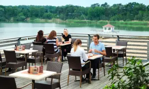 Gemütliches Restaurant am Wasser mit Gästen, die Kaffee und Getränke genießen. Im Hintergrund ein ruhiger See und grüne Bäume.