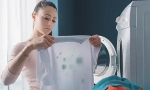 Zmartwiona kobieta trzyma białą koszulkę z zielonymi plamami, widoczne odbarwione ubrania po praniu.