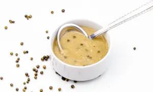 Aksamitny sos z zielonego pieprzu w białej miseczce, z łyżką zanurzoną w sosie. Wokół rozsypane ziarna zielonego pieprzu.