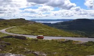 Eine malerische England Landschaft mit einer kurvigen Straße, einem roten Campervan und einem See in der Ferne unter bewölktem Himmel.