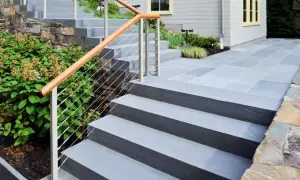 Escalier extérieur en béton, recouvert de dalles grises, avec main courante en bois et câbles. Un chemin en pierre mène à la maison.