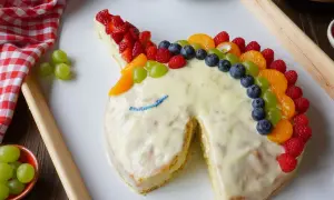 Ein fantasievoller Kuchen ohne Zucker in Einhornform, verziert mit bunten Früchten wie Erdbeeren, Blaubeeren und Mandarinen.