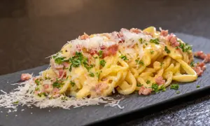 Eine Portion Spaghetti Carbonara mit Speckwürfeln, Petersilie und Parmesan auf einem dunklen Teller.